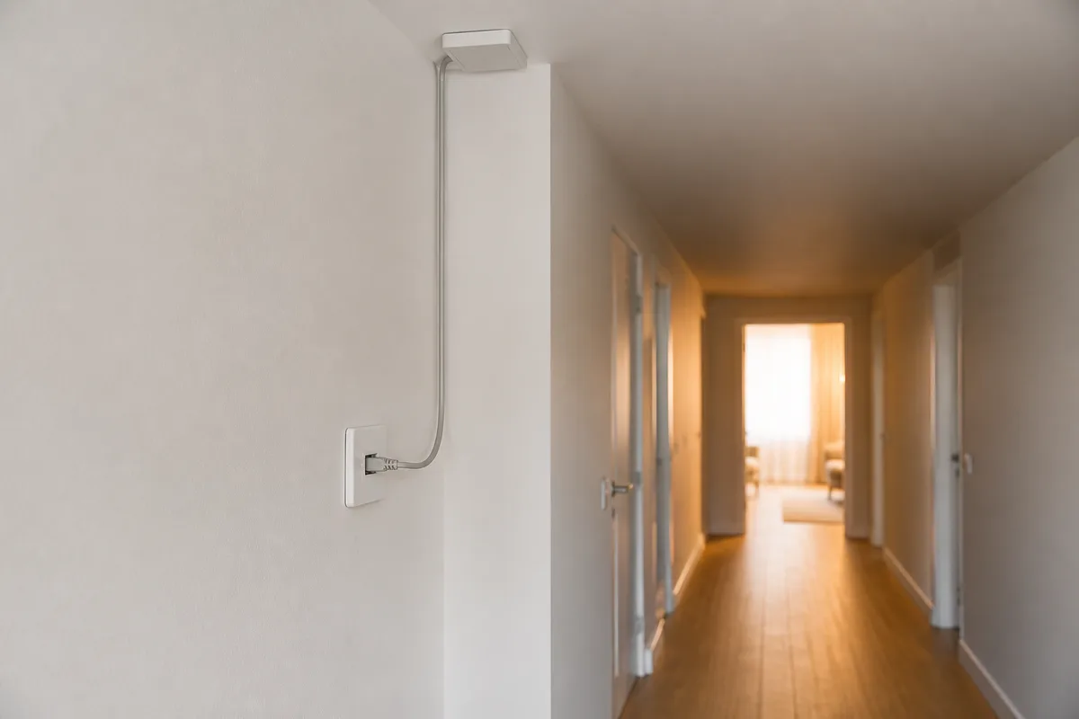 Prise réseau murale RJ45 posée dans un couloir clair, câble raccordé vers un boîtier au plafond